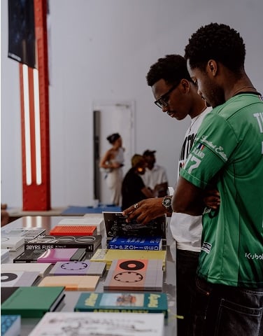 The image shows two men standing side by side, closely examining a table filled with various books and notebooks. The background features a few other people engaged in conversation or activities within an indoor setting.