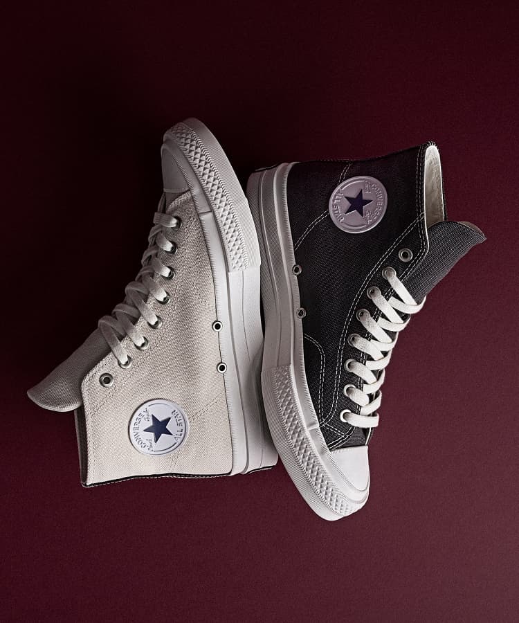 The image shows a pair of white Converse First String high-top sneakers placed on a black leather seat. One shoe is upright, displaying the embossed Converse logo, while the other lies flat, showing its black sole with a yellow Vibram label.