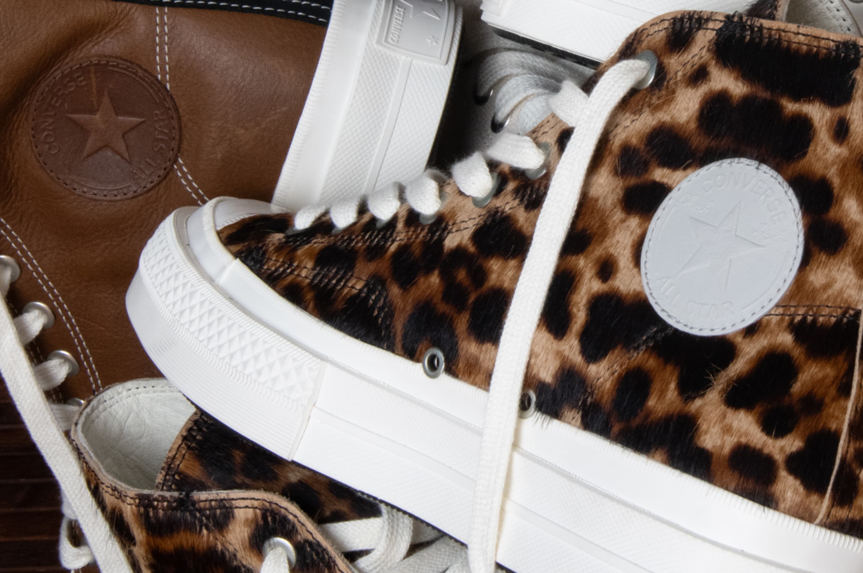 The image shows close-up views of white Converse sneakers, highlighting different styles and details like stitching, laces, and logos. The left side features a high-top with brown accents, while the right side focuses on low-top shoes with textured rubber soles.