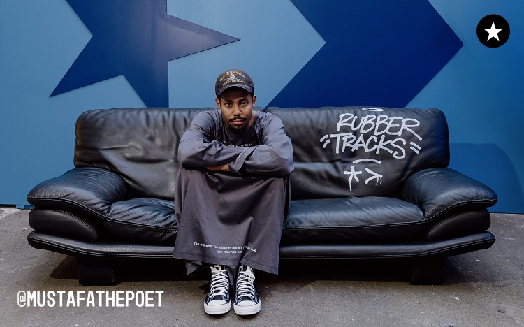 The image shows a person sitting cross-armed on a black leather couch with the words &ldquo;RUBBER TRACKS&rdquo; written on it, set against a blue wall featuring large star shapes. The individual is wearing dark clothing, sneakers, and a cap, with the Instagram handle &ldquo;@MUSTAFATHEPOET&rdquo; visible in the bottom left corner.