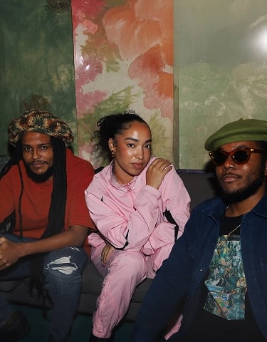 The image shows three people sitting closely together on a couch against a colorful, floral-patterned wall. They are dressed casually, with one person wearing a pink outfit in the center, while the others wear darker clothing and hats.