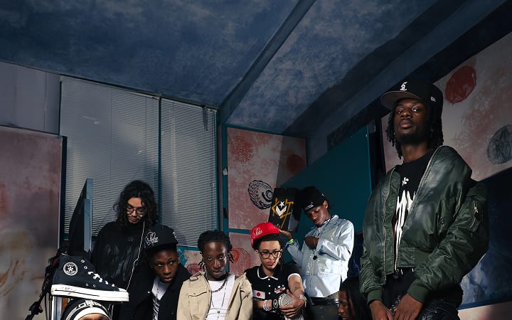 The image shows a group of seven people posing indoors against a backdrop with abstract artwork and blue walls. They are dressed in casual streetwear, including jackets, hats, and sneakers, with one person prominently holding up a sneaker.