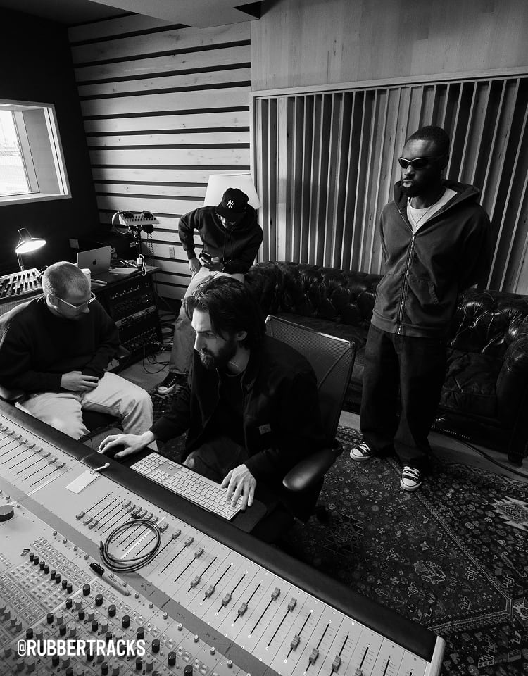 The image shows four people in a music studio, with one person seated at a mixing console operating equipment while the others stand or sit nearby. The room features soundproofing panels on the walls and various audio gear, creating a professional recording environment.