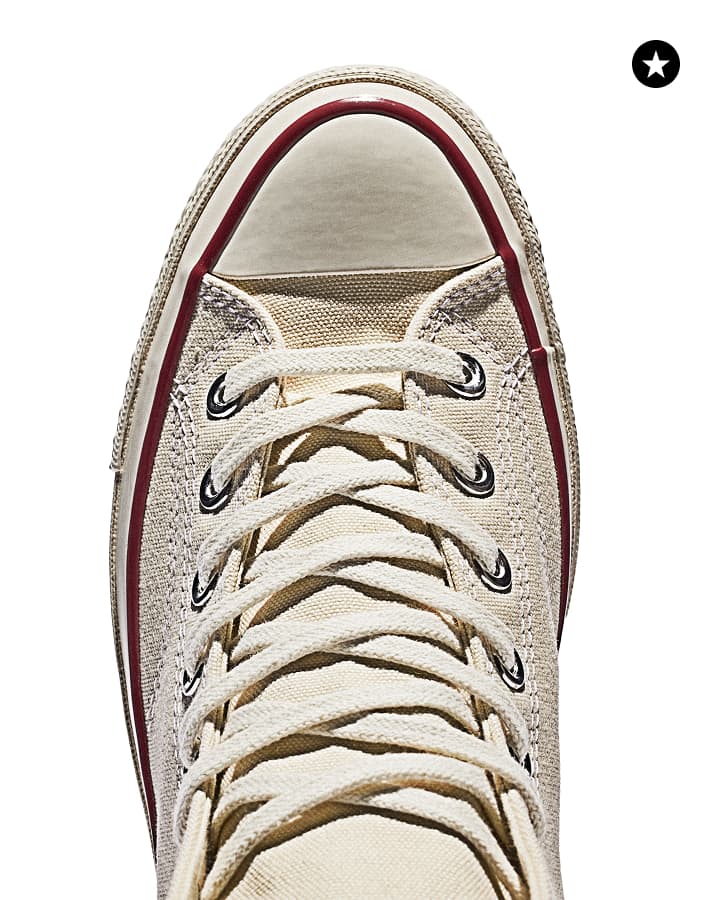 The image shows a pair of classic Converse Chuck Taylor All Star sneakers in an off-white or beige color. One shoe is positioned lying flat on its sole, showing the top view with laces neatly tied, while the other shoe is balanced horizontally on top of it, displaying a side profile. 