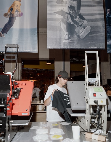 The image shows a person standing between two large heat press machines, holding a black t-shirt. Above them are two large posters featuring skateboarders in action. The setting appears to be an indoor event or workshop with people and equipment visible in the background.