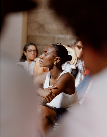 The image shows a group of people seated indoors, with the focus on a woman in the center wearing a white sleeveless top and gold earrings. The background is softly blurred, highlighting the central figure who appears to be attentively listening or engaged in an event.