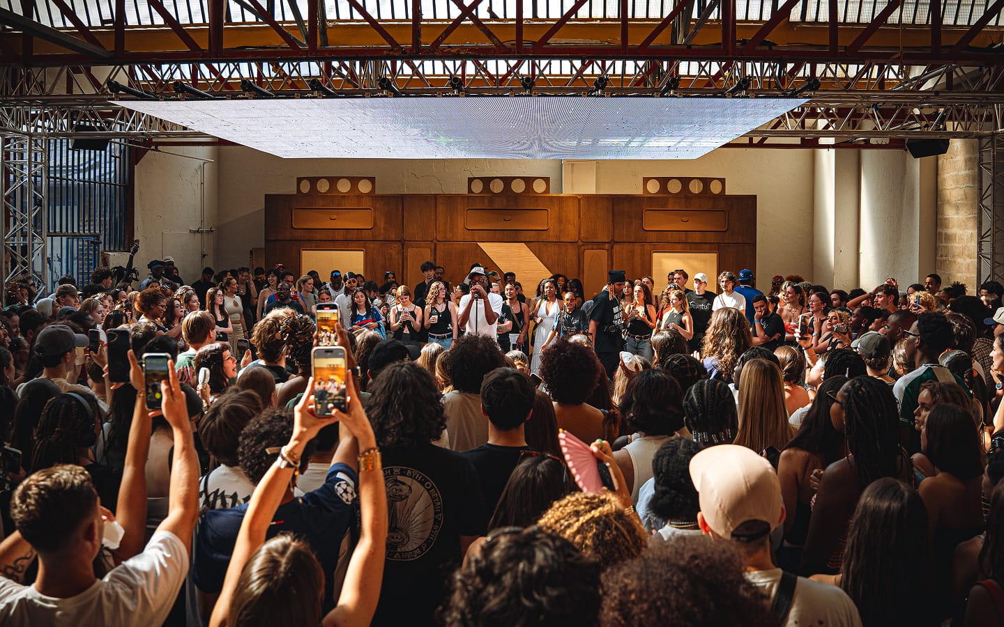 The image shows a large crowd of people gathered indoors, many holding up their phones to record or take pictures of a group of individuals standing on a stage or platform. The setting appears to be an event or performance in a spacious room with industrial-style architecture and wooden paneling in the background.