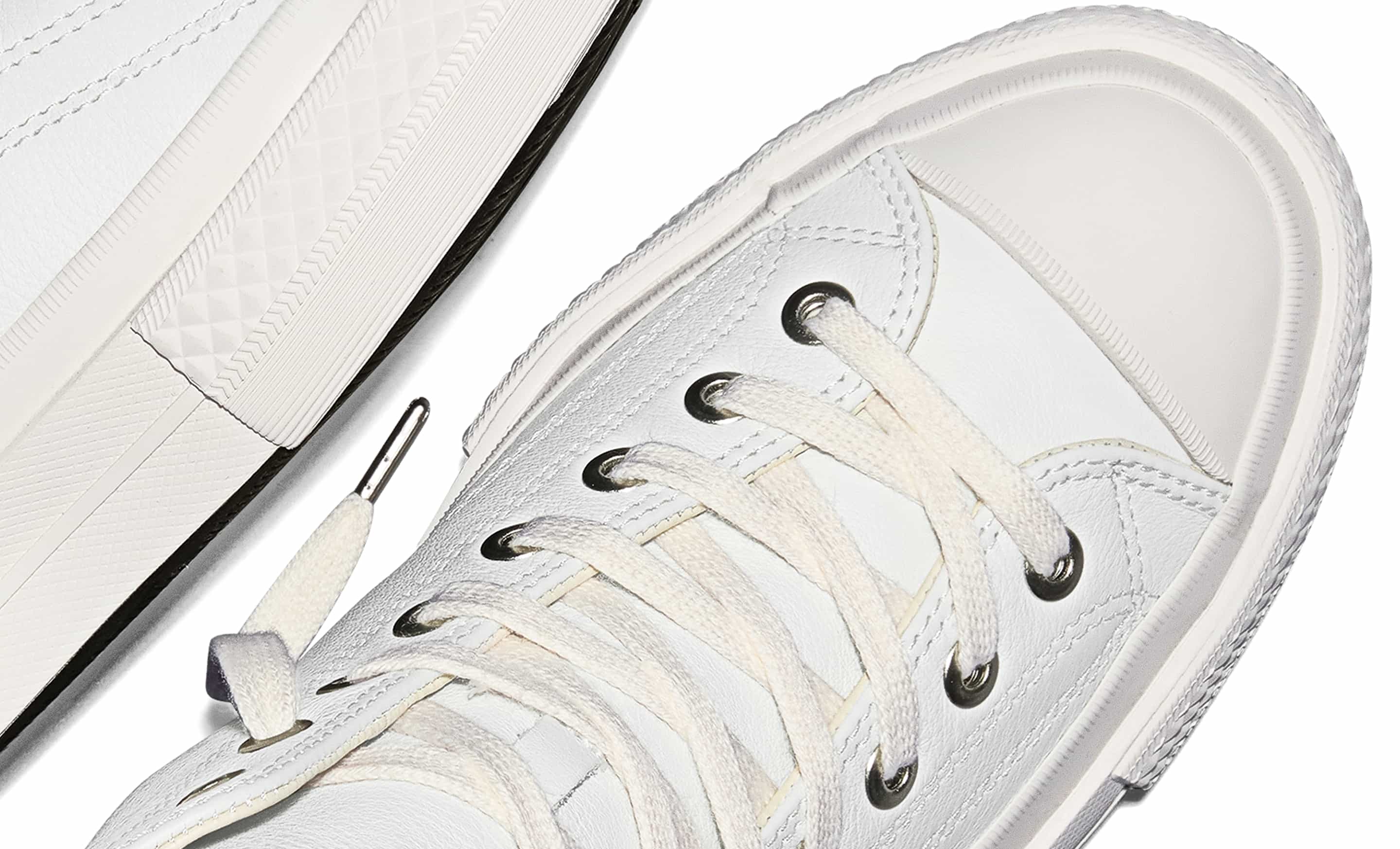 The image shows a close-up of a white sneaker with white laces threaded through metal eyelets. The shoe features a smooth rubber toe cap and detailed stitching along the sides.