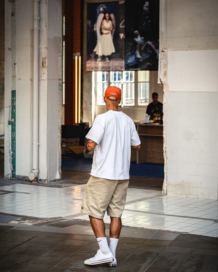 The image shows a man standing indoors with his back to the camera, wearing a white t-shirt, beige shorts, white socks, white sneakers, and an orange cap. In the background, there are two large posters featuring people and a person behind a counter in a well-lit room.