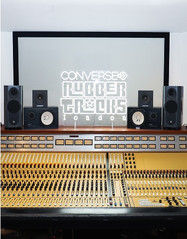 The image shows a professional audio mixing console with multiple speakers arranged on top of it. Behind the equipment, there is a large screen displaying the illuminated text &ldquo;CONVERSE RUBBER TRACKS LONDON.&rdquo;