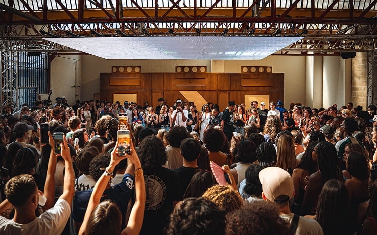 The image shows a large crowd of people gathered indoors, many holding up their phones to record or take pictures. At the front, a group of individuals appears to be performing or speaking on a stage under a metal framework with overhead lighting.