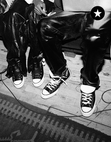 The black-and-white image shows two people sitting side by side, both wearing shiny leather pants and Converse-style sneakers. The focus is on their legs and feet, with one person holding a laptop on their lap.