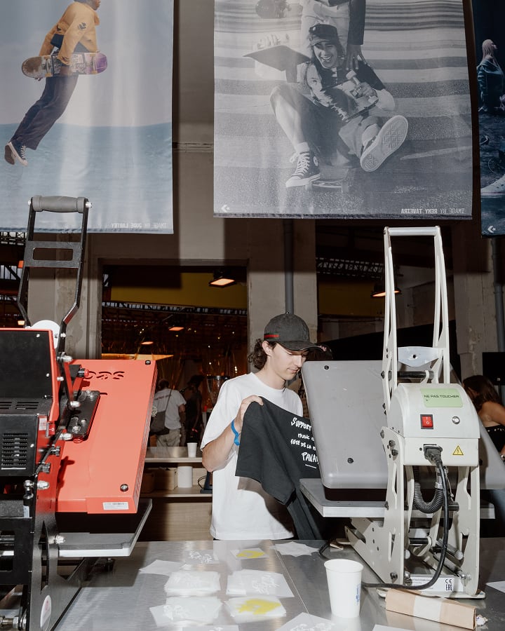 The image shows a person holding a black T-shirt in what appears to be a printing or merchandise booth with heat press machines on the table. Above them, there are large posters featuring skateboarding images hanging from the ceiling.