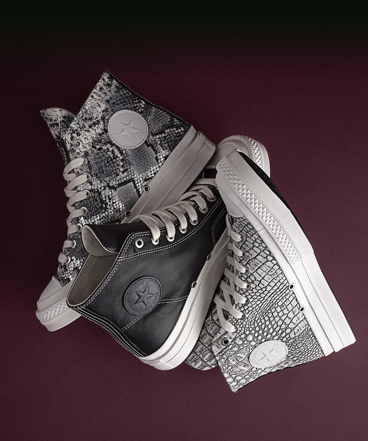 The image shows a pair of white Converse First String high-top sneakers placed on a black leather seat. One shoe is upright, displaying the embossed Converse logo, while the other lies flat, showing its black sole with a yellow Vibram label.