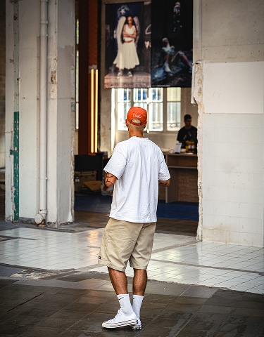 The image shows a man wearing a white t-shirt, beige shorts, white socks, and sneakers standing indoors with his back to the camera. He is looking towards an area with large windows and some artwork or posters hanging on the wall.