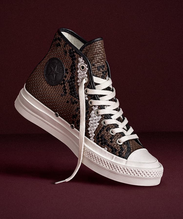 The image shows a pair of white Converse First String high-top sneakers placed on a black leather seat. One shoe is upright, displaying the embossed Converse logo, while the other lies flat, showing its black sole with a yellow Vibram label.