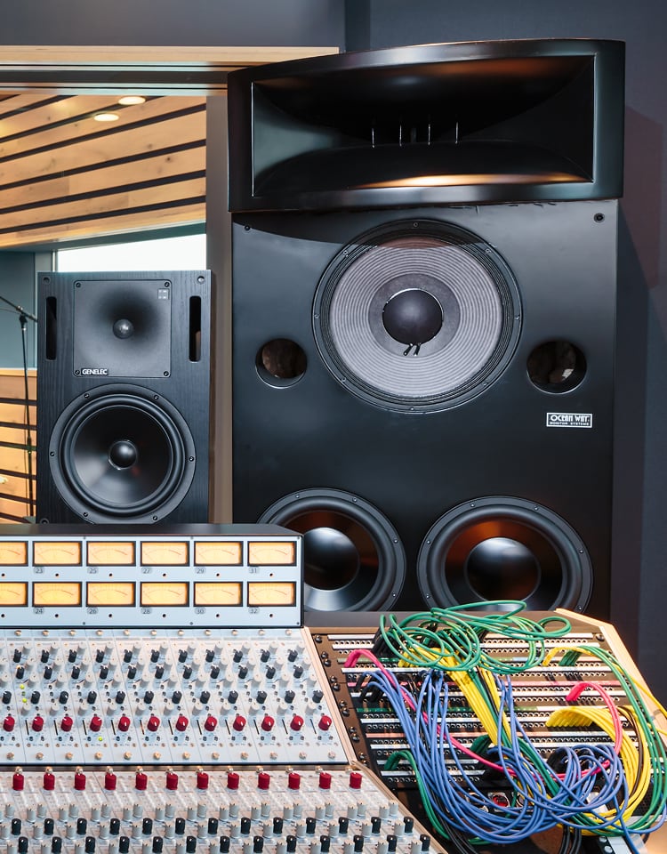 The image shows a professional audio mixing console with numerous knobs, switches, and patch cables in the foreground, set in front of two large studio monitor speakers. The setting appears to be a recording or sound engineering studio with acoustic paneling visible through a window on the left.