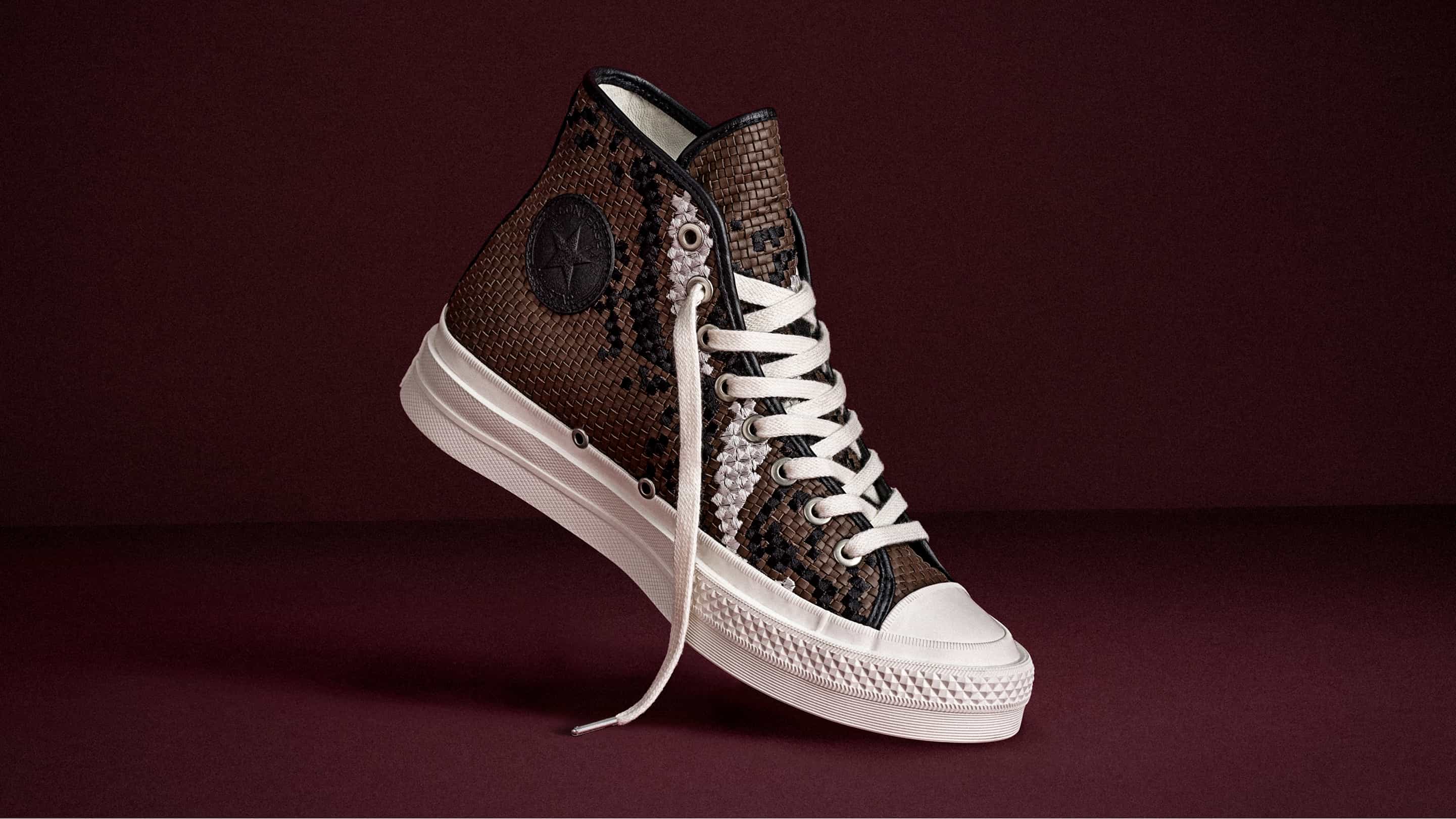 The image shows a pair of white Converse First String high-top sneakers placed on a black leather seat. One shoe is upright, displaying the embossed Converse logo, while the other lies flat, showing its black sole with a yellow Vibram label.