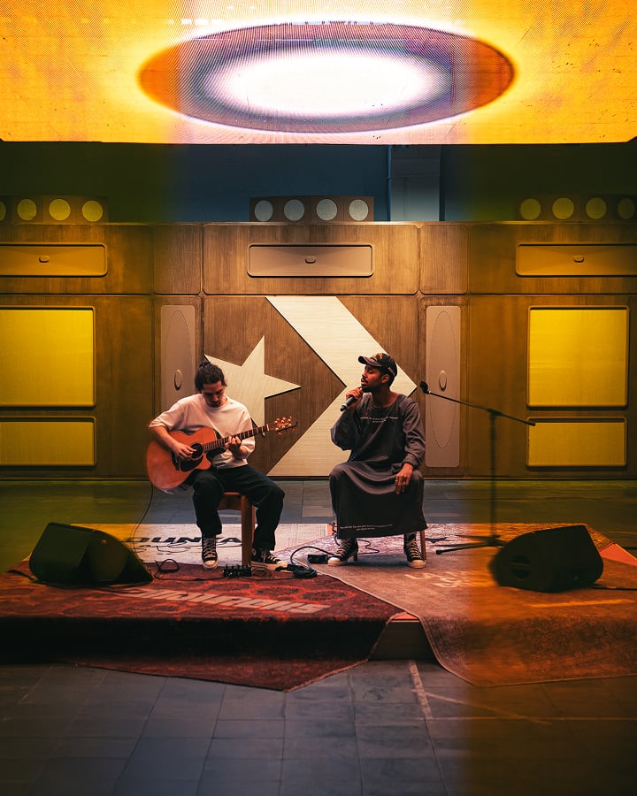 The image shows two people performing on a small stage, with one playing an acoustic guitar and the other singing into a microphone. The background features a wooden wall with geometric designs, and warm lighting creates an intimate atmosphere.