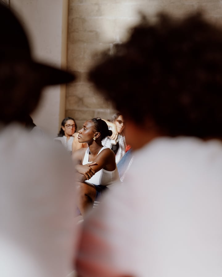 The image shows a group of people sitting indoors, with the focus on a woman in a white tank top and sunglasses. The foreground is blurred, featuring two individuals whose faces are not visible clearly.