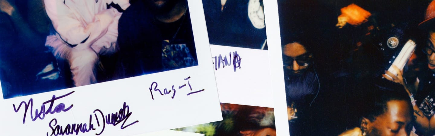 The image shows a close-up of several overlapping Polaroid photos with handwritten signatures and notes on the white borders. The background is dark, featuring people in casual attire, one holding a rolled-up item, possibly a poster or paper.