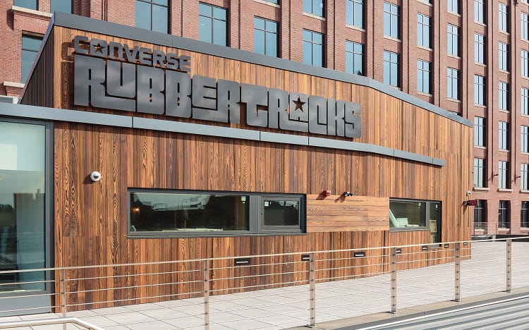 The image shows a modern wooden building facade with large black signage that reads &ldquo;CONVERSE RUBBERTRACKS.&rdquo; The structure is set against a backdrop of a taller brick building, and there is a metal railing in front of the building.