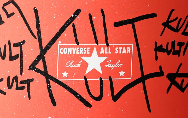 The image shows a red background with the white Converse All Star Chuck Taylor logo prominently displayed in the center. Surrounding the logo are large, black graffiti-style letters spelling out &ldquo;KULT&rdquo; multiple times.