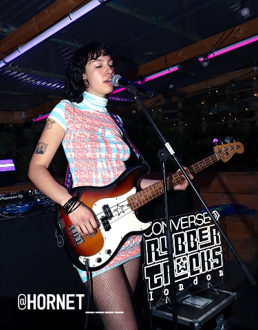 The image shows a person playing an electric bass guitar and singing into a microphone on stage, wearing a patterned dress and fishnet stockings. The background includes musical equipment and a sign that reads &ldquo;CONVERSE RUBBER TRACKS LONDON.&rdquo;
