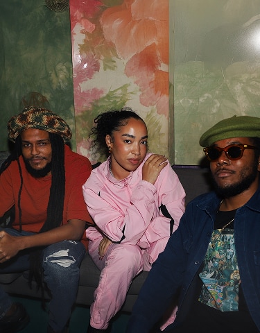 The image shows three people sitting closely together on a couch against a colorful, floral-patterned wall. They are dressed casually, with one person wearing a pink outfit in the center, while the others wear darker clothing and hats.