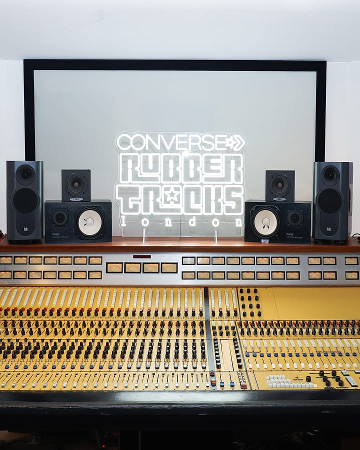 The image shows a professional audio mixing console with multiple knobs, sliders, and meters in the foreground. Behind it is a neon sign that reads &ldquo;CONVERSE RUBBER TRACKS london,&rdquo; flanked by several studio monitor speakers.