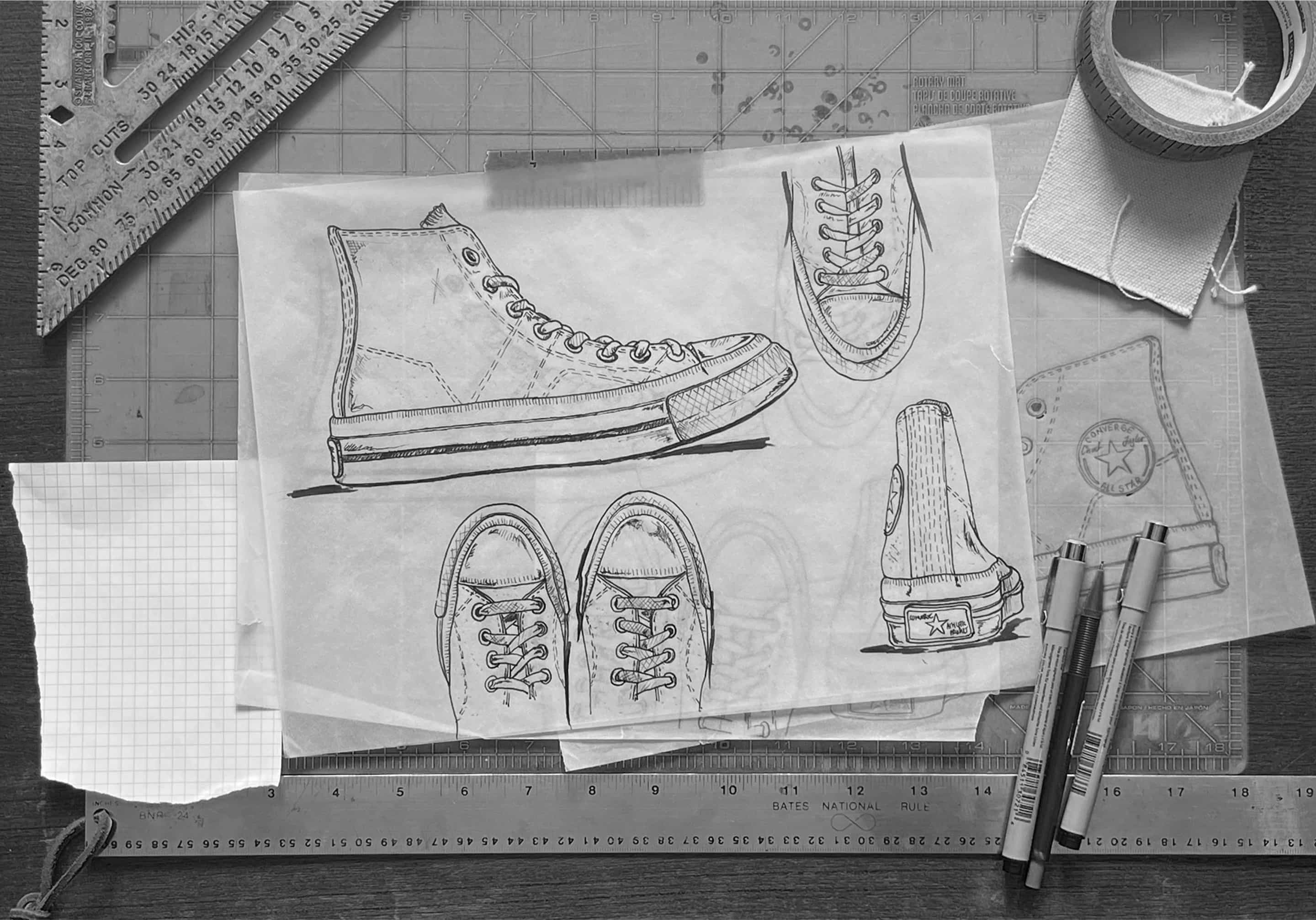 This image shows a designer’s workspace featuring detailed hand-drawn sketches of the Converse First String shoe, including side, top, front, and rear views on translucent tracing paper. Surrounding the sketches are drafting tools such as metal rulers, fine-tip pens, masking tape on fabric swatches, and graph paper, highlighting a precise and creative footwear design process.