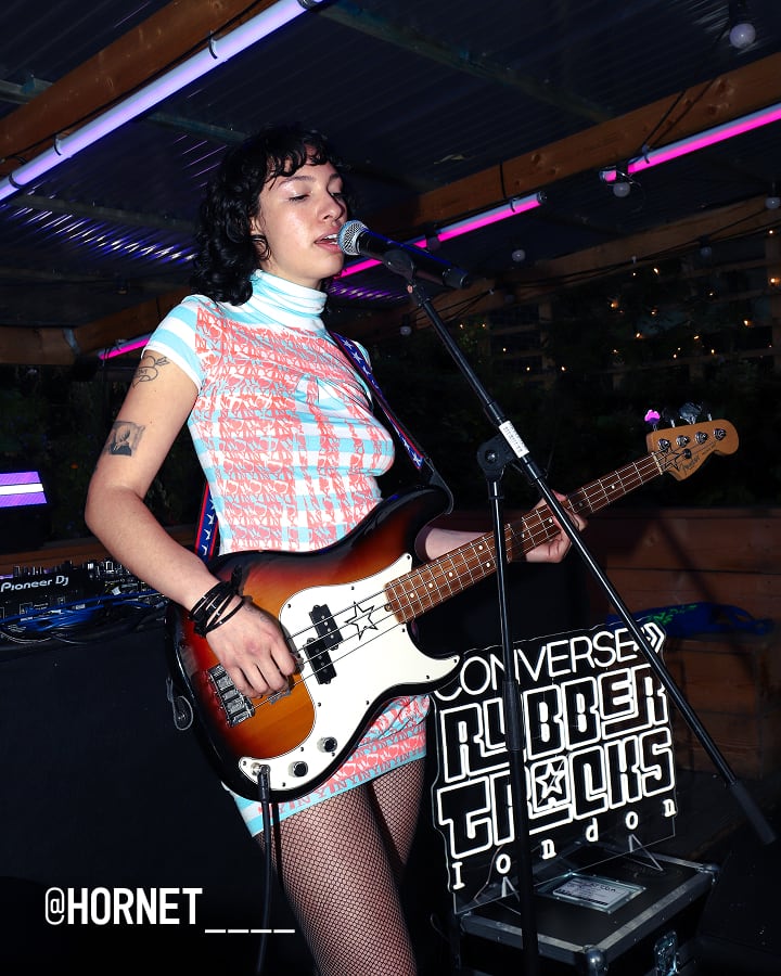 The image shows a person playing an electric bass guitar on stage, standing in front of a microphone. The background features neon lights and a sign that reads &ldquo;CONVERSE RUBBER TRACKS LONDON.&rdquo;