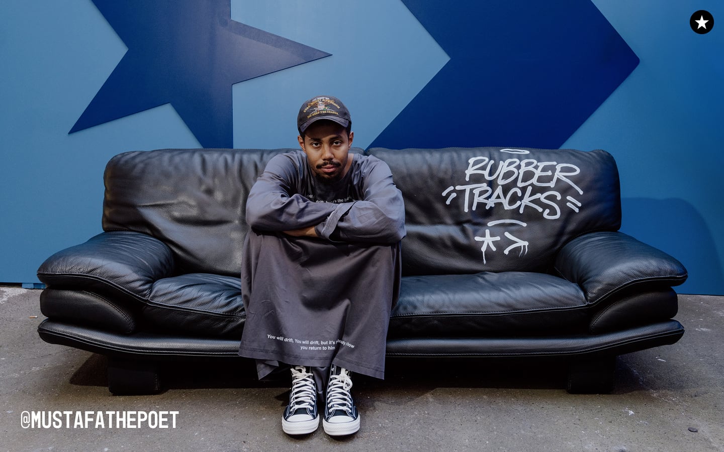 The image shows a person sitting cross-armed on a black leather couch with the words &ldquo;RUBBER TRACKS&rdquo; written in white graffiti-style text on the right side of the couch. The background features a blue wall with large star and arrow shapes, and the Instagram handle &ldquo;@MUSTAFATHEPOET&rdquo; is visible in the bottom left corner.