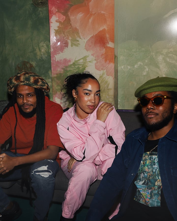 The image shows three people sitting on a couch against a colorful, floral-patterned wall. They are dressed casually, with one person wearing a pink tracksuit, another in a red shirt and knit hat, and the third in a dark jacket and green beret.