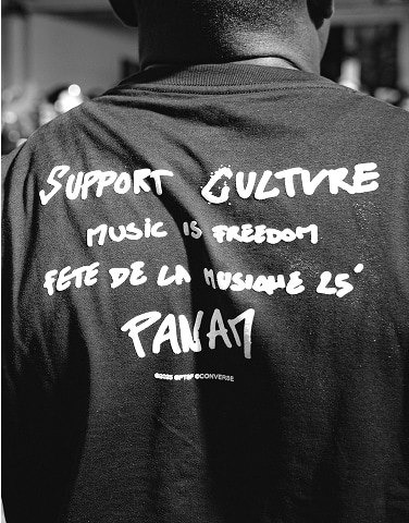 The image shows the back of a person wearing a black shirt with white text that reads &ldquo;SUPPORT CULTURE,&rdquo; &ldquo;MUSIC IS FREEDOM,&rdquo; &ldquo;FETE DE LA MUSIQUE 25&deg;,&rdquo; and &ldquo;PANAM.&rdquo; The photo is in black and white, focusing on the shirt&rsquo;s message.