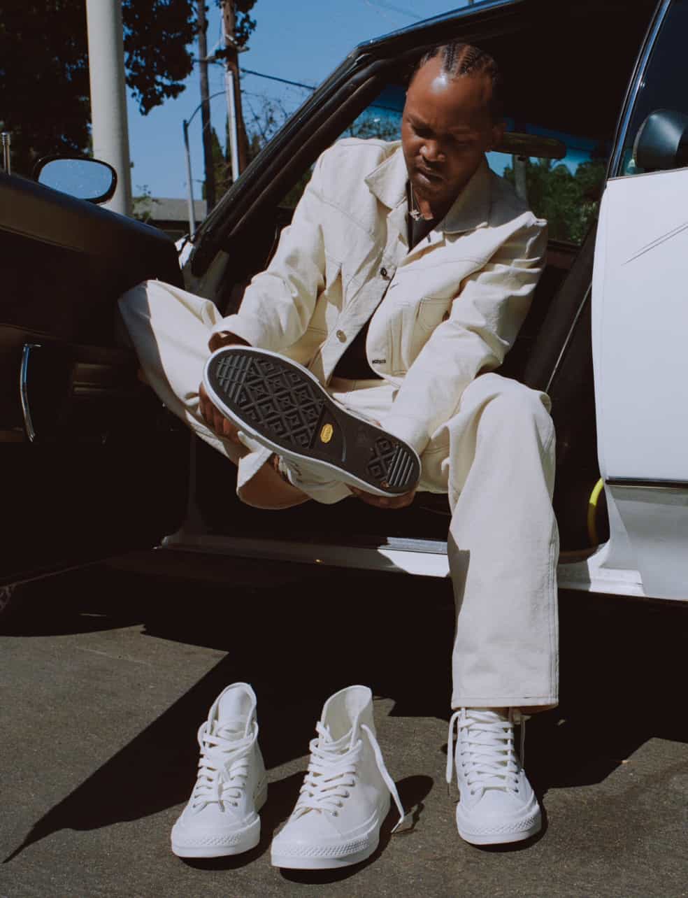 A person dressed in cream-colored clothing sits in the open doorway of a white car, holding up the sole of one shoe towards the camera. Two white high-top sneakers are placed on the ground in front of them, with one shoe worn on their foot.