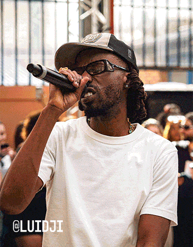 The image shows a person wearing a white t-shirt and a black-and-white cap, holding a microphone close to their mouth as if performing or speaking. The background features an outdoor setting with several people watching the individual.