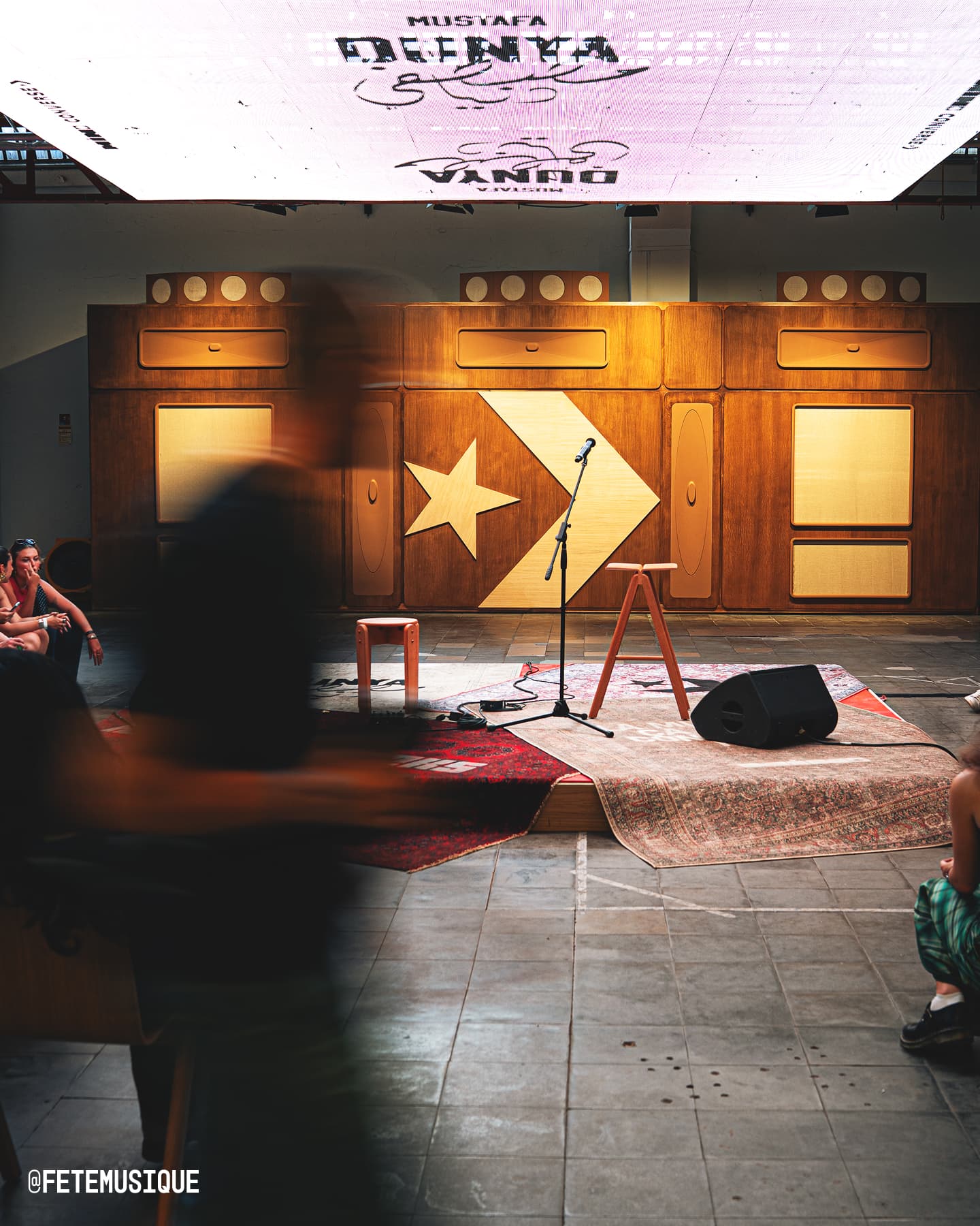 The image shows a small stage setup with a microphone stand, two stools, and a speaker on rugs in front of a wooden wall decorated with geometric shapes including a star. There are blurred figures moving in the foreground and some seated people on the sides, suggesting an intimate live performance or event setting.