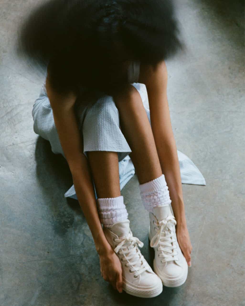 A person is sitting on the floor with their legs bent and arms reaching toward their feet, wearing white sneakers and socks. Their head is blurred in motion, obscuring their face.