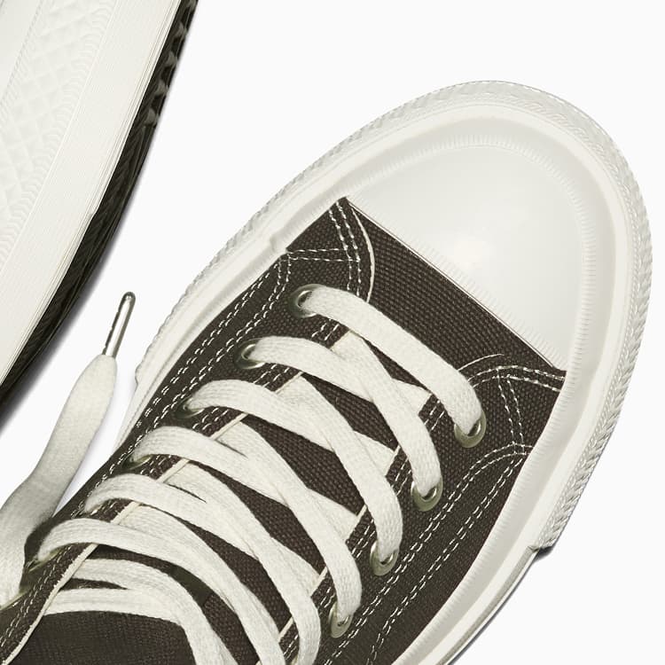 The image shows a close-up of a white sneaker with white laces threaded through metal eyelets. The shoe features a smooth rubber toe cap and detailed stitching along the sides.