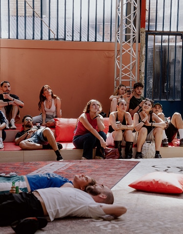 The image shows a group of people sitting and lounging on red cushions and benches in an indoor space with large windows and industrial-style metal beams. In the foreground, two individuals are lying down on a patterned rug, appearing relaxed or resting.