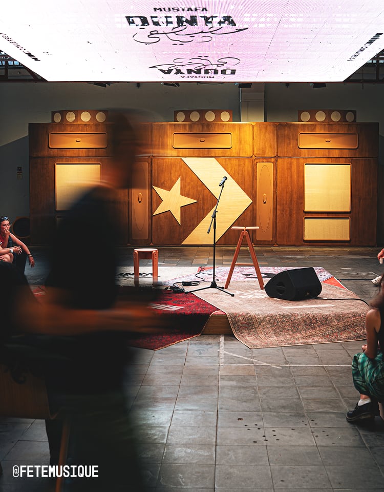 The image shows a small stage setup with a microphone stand, two stools, and a speaker on rugs in front of a large wooden wall featuring the Converse logo (a star and chevron). There are blurred people moving around the area, suggesting an informal or intimate event setting.