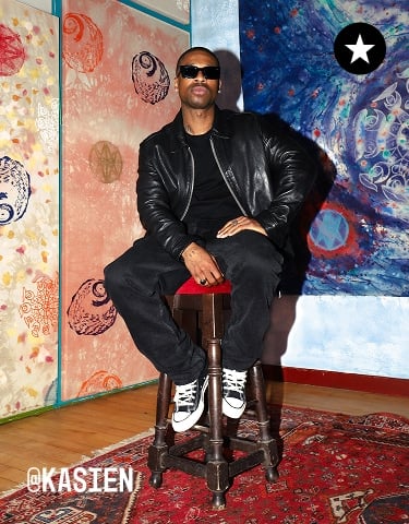 The image shows a person sitting on a wooden stool against a colorful backdrop featuring abstract and cosmic-themed artwork. The individual is dressed in black clothing with white sneakers, and the setting includes a patterned red rug on the floor.