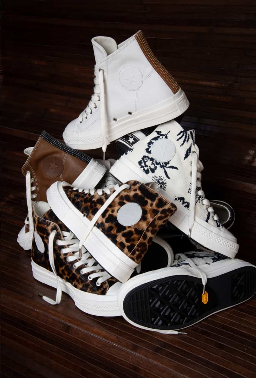 A pile of high-top sneakers with various designs, including white, brown leather, floral, and leopard print patterns, is stacked on a dark wooden surface. All the shoes have white soles and laces, showcasing different textures and colors.