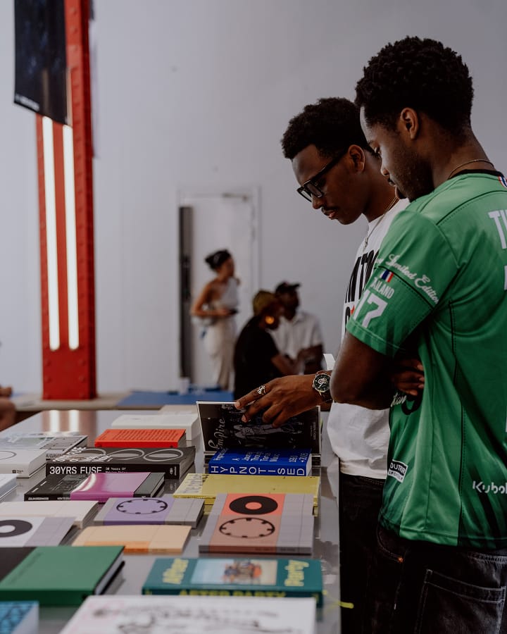 The image shows two men standing at a table filled with various books, with one of them flipping through a book. In the background, three people are sitting and standing near a white wall in a casual setting.