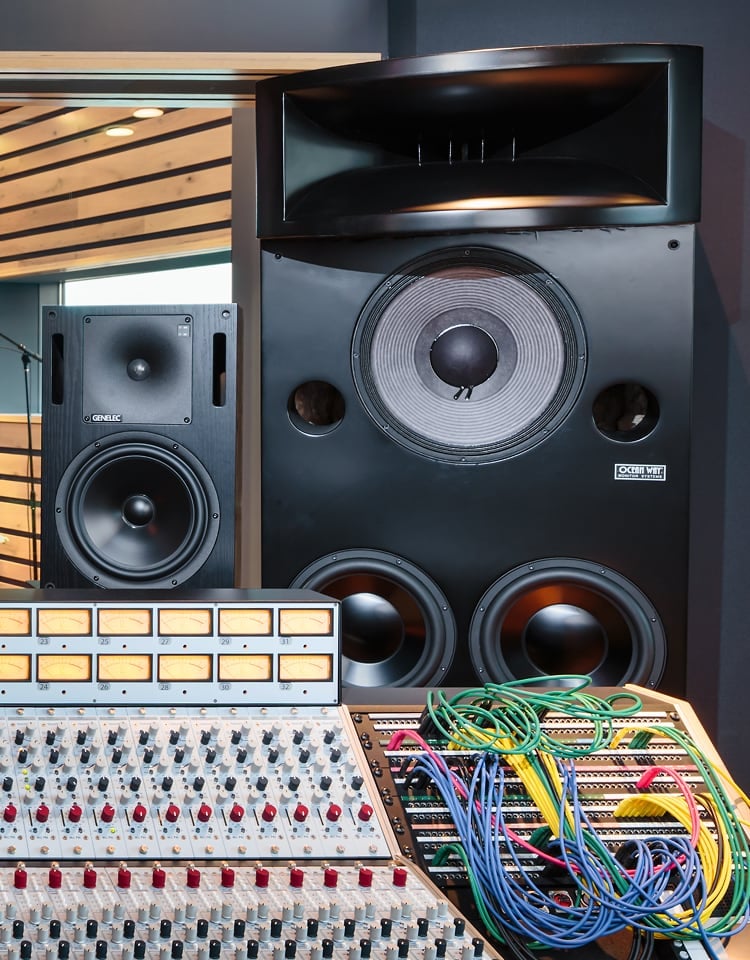 The image shows a professional audio mixing console with numerous knobs, switches, and patch cables in the foreground, set in front of two large studio monitor speakers. The setting appears to be a recording or sound engineering studio with acoustic paneling visible through a window on the left.