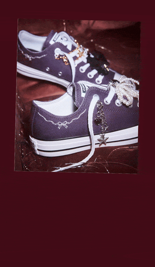 A pair of purple low-top canvas sneakers rest on a glossy burgundy surface. They’re decorated with pearl strands, metallic charms, and delicate white stitching details.