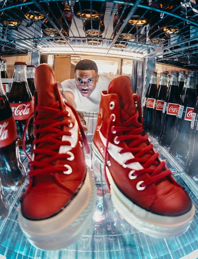 The image shows a person reaching into a refrigerator stocked with glass bottles of Coca-Cola and a pair of red high-top sneakers placed inside. The perspective is from inside the fridge looking outward toward the person.