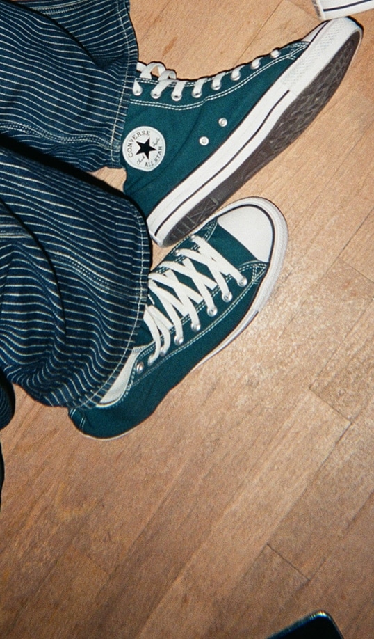 The image shows a person wearing teal Converse All Star high-top sneakers paired with blue and white striped pants. The person is standing on a wooden floor, and the photo is taken from above.