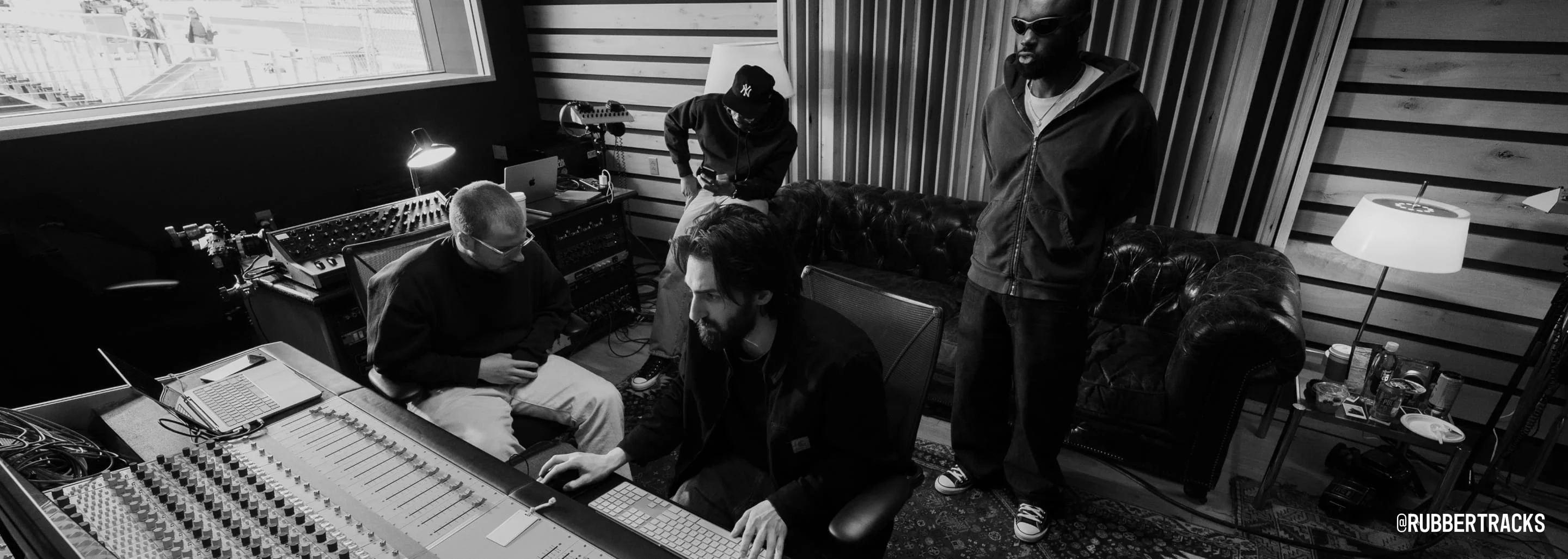 The image shows a black-and-white scene of four people in a music studio, with one person seated at a mixing console and others either sitting or standing nearby. The room features soundproofing panels, various audio equipment, and a window letting in natural light, with the handle @RUBBERTRACKS visible in the bottom right corner.